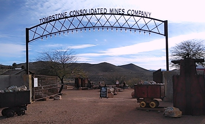 Entrance to the Tombstone Consolidated Mines Company with a metal archway, mining carts filled with rocks, and a desert landscape with hills and trees in the background.