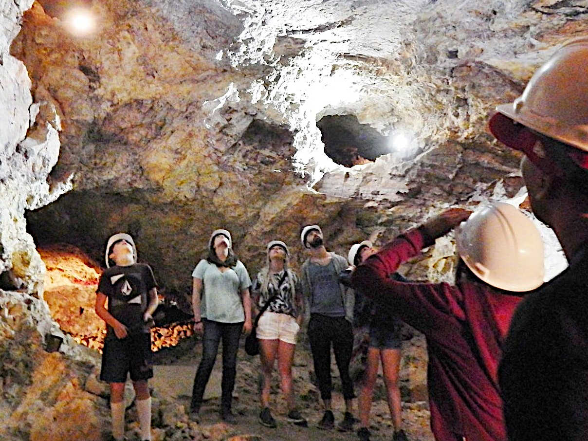 Group of visitors wearing hard hats inside a well-lit underground mine, looking upwards at the rocky ceiling and illuminated openings.