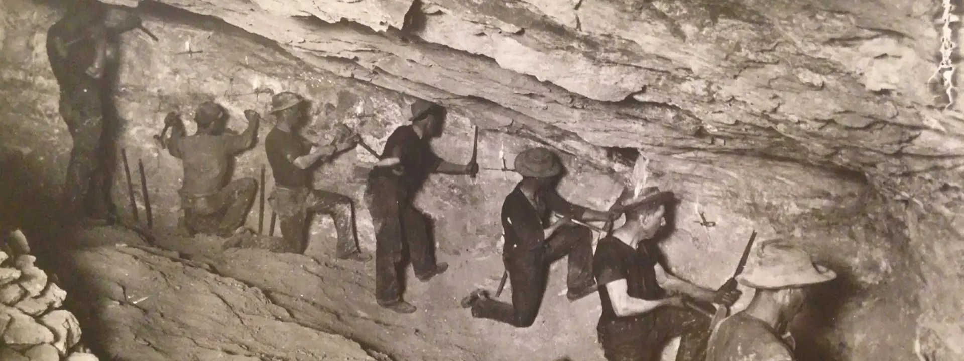 Historic black-and-white photograph of miners working underground, using pickaxes and tools to extract ore from rock walls in a dimly lit tunnel.