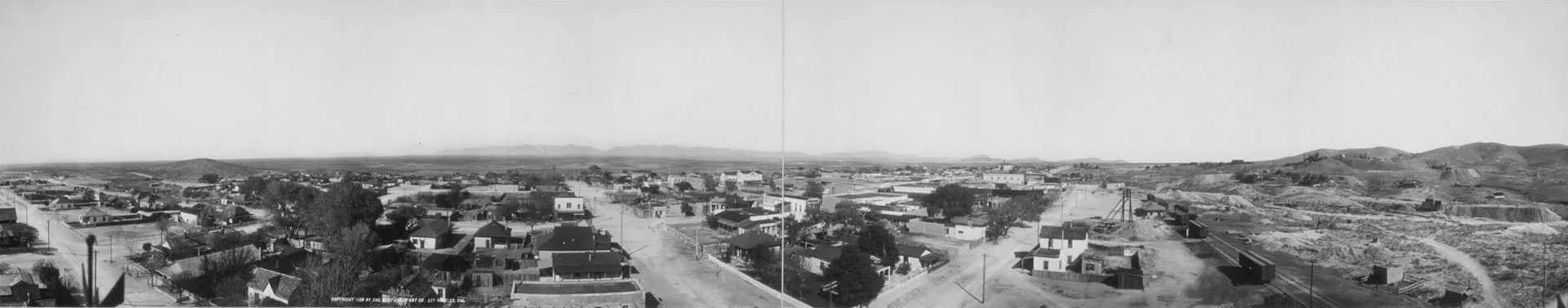 Panoramic black-and-white photograph of Tombstone, Arizona, featuring a historic view of buildings, streets, and surrounding desert landscape with distant hills under a clear sky.