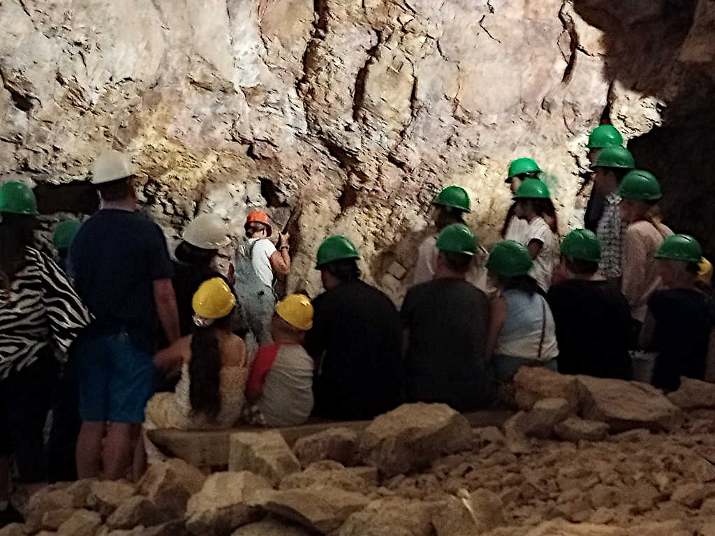 Group of visitors wearing green, yellow, and white hard hats gathered around a tour guide demonstrating mining techniques inside an underground mine.