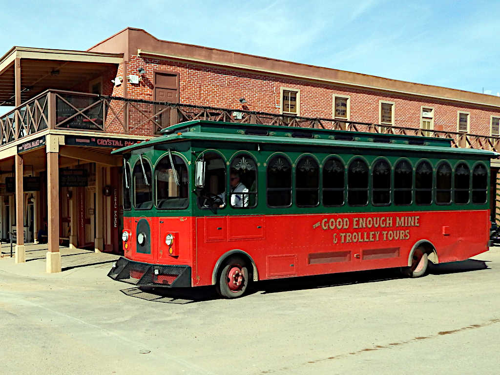 Red and green trolley labeled 'Good Enough Mine & Trolley Tours' passing in front of the historic Crystal Palace building with a brick facade and wooden balcony.