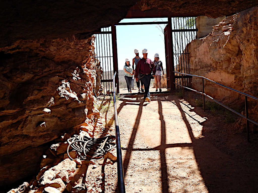 Visitors wearing hard hats entering a mine tunnel with rocky walls, sunlight streaming through the gate, and safety railings on both sides of the path.