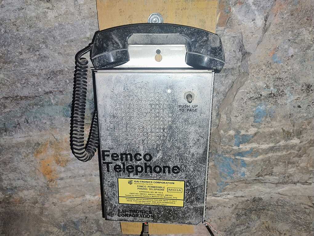 Wall-mounted emergency Femco telephone inside a mine, with a metal casing and a coiled cord, against a rough rock wall.