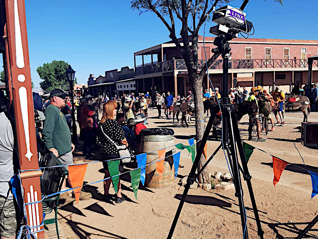 Event setup on a dirt street in a western-style town, with colorful bunting, a tripod-mounted camera labeled 'LYNX,' and a crowd of people and donkeys in the background.