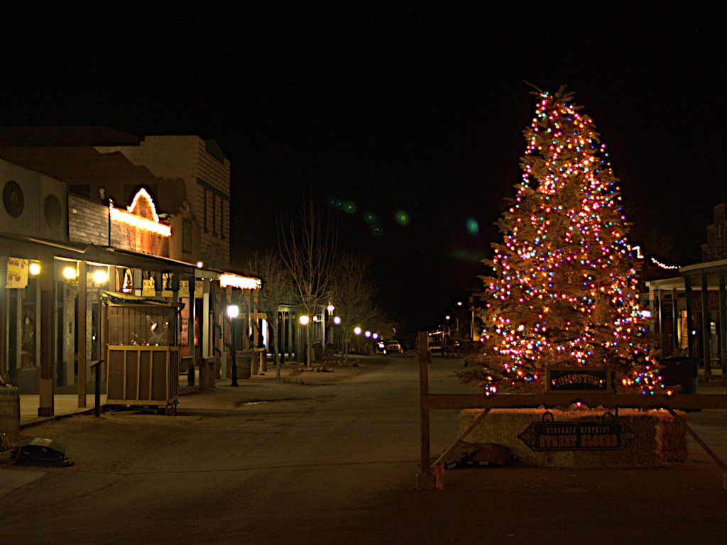 Nighttime view of Tombstone's historic district featuring a large Christmas tree adorned with colorful lights, surrounded by old western-style buildings illuminated with festive decorations.