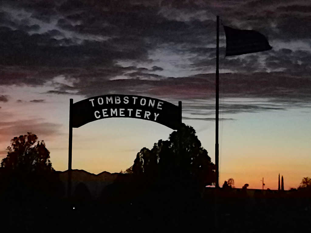 Silhouette of the Tombstone Cemetery entrance sign and a flagpole against a dramatic sunset with dark clouds and colorful twilight hues.