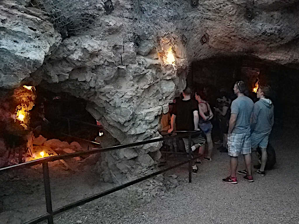 Visitors gathered inside an underground mine illuminated by warm lights, observing rock formations and a narrow passage with safety railings.