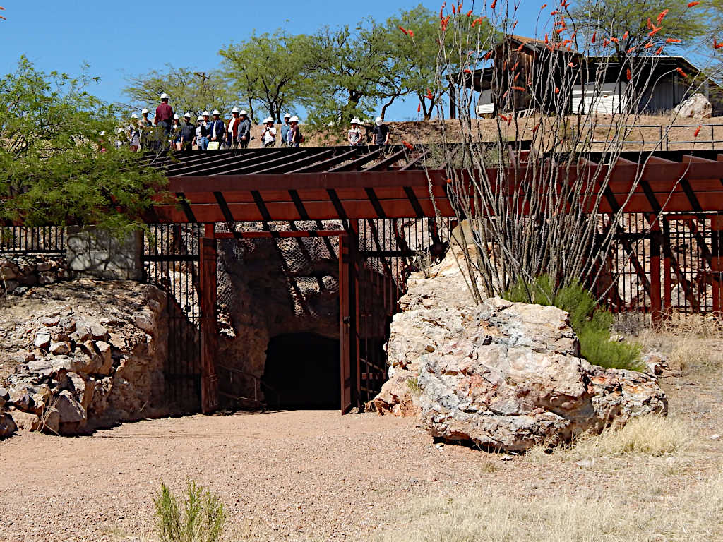 Entrance to an underground mine surrounded by rocky terrain and desert vegetation, with a group of visitors in hard hats visible on the structure above.