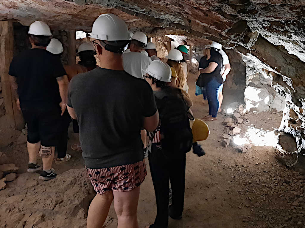 Group of visitors wearing hard hats walking through a narrow underground mine tunnel with exposed rock walls and wooden supports.