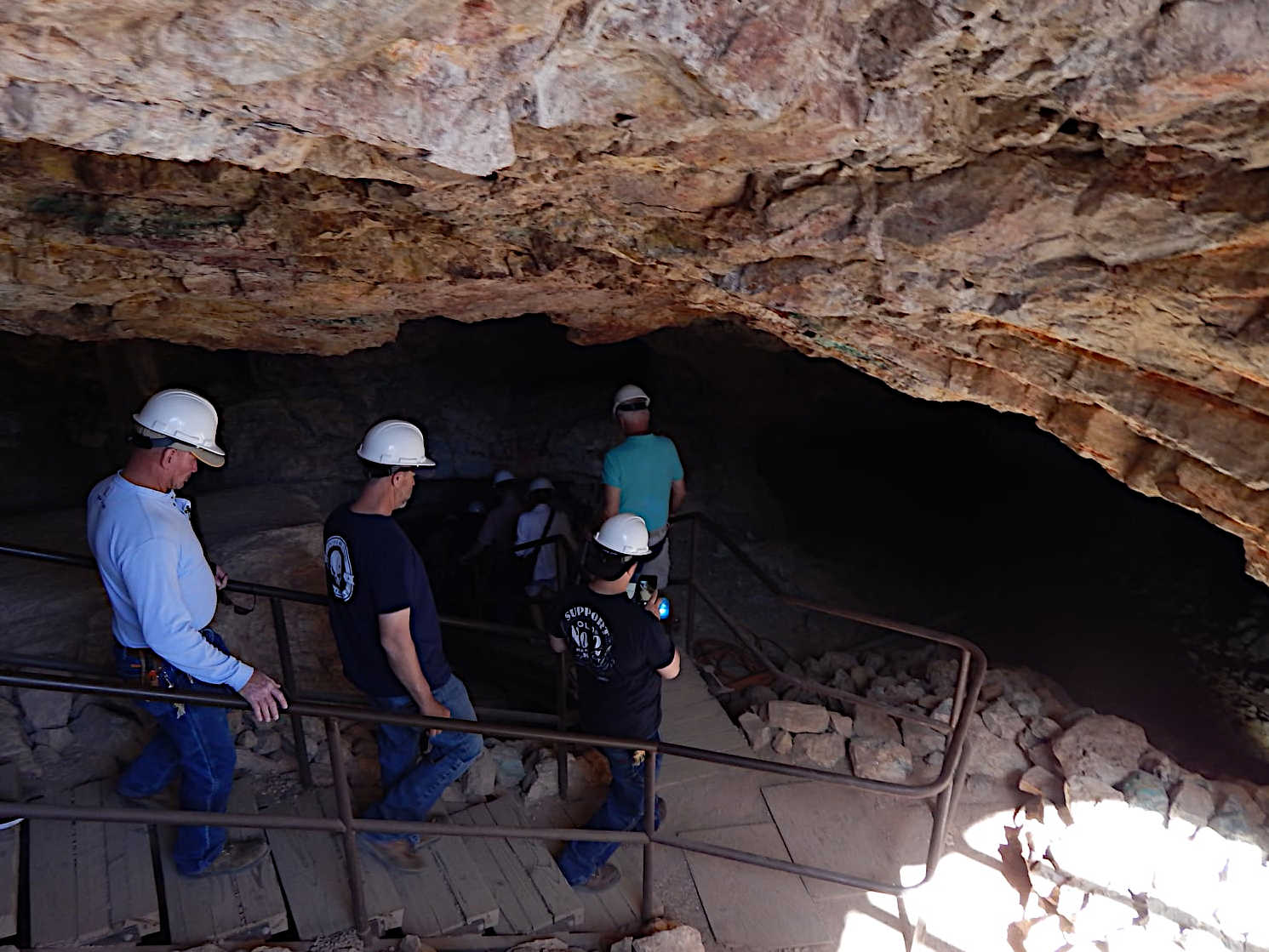 Entrance to an underground mine with a wooden walkway and metal handrails, surrounded by large rocks and rugged stone walls.