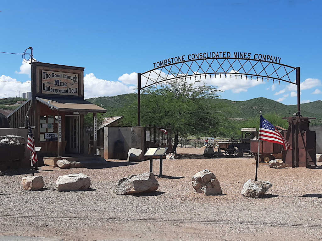 Entrance to the Good Enough Mine Underground Tour in Tombstone, Arizona, featuring a rustic wooden building, a metal archway reading 'Tombstone Consolidated Mines Company,' American flags, old mining equipment, and desert landscape in the background.