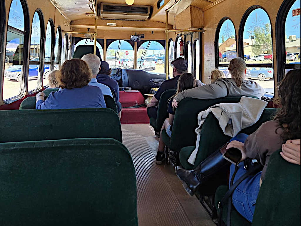 Interior of a trolley bus with green upholstered seats and passengers looking forward during a daytime tour. The view through the windows shows a small-town street with parked cars and buildings.