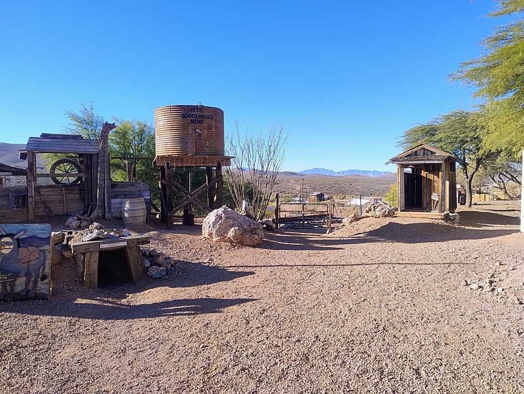 Outdoor area at the Good Enough Mine in Tombstone, Arizona, featuring historic mining structures including a water tank labeled 'Good Enough Mine,' rustic wooden buildings, and a desert landscape with distant mountains under a clear blue sky.