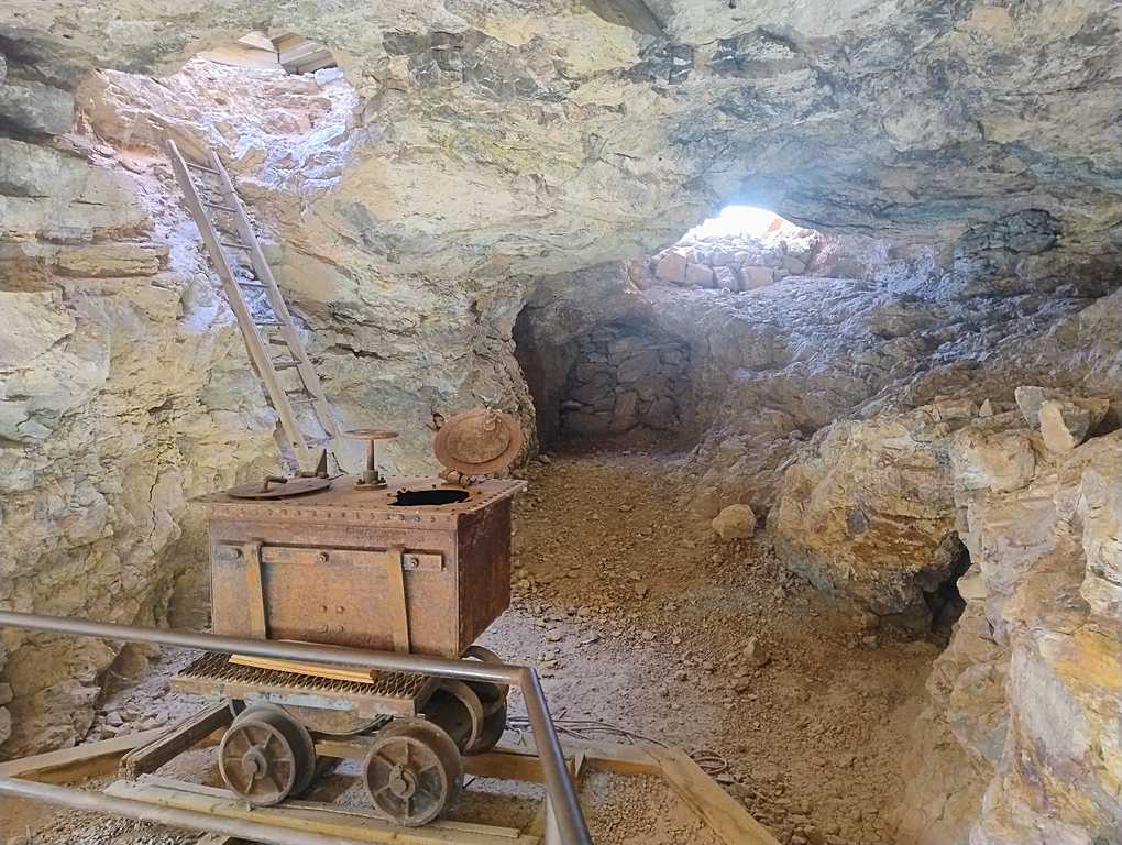 Underground mine chamber with a rusty ore cart on rails, a wooden ladder leading to an opening, and rocky walls illuminated by natural light.