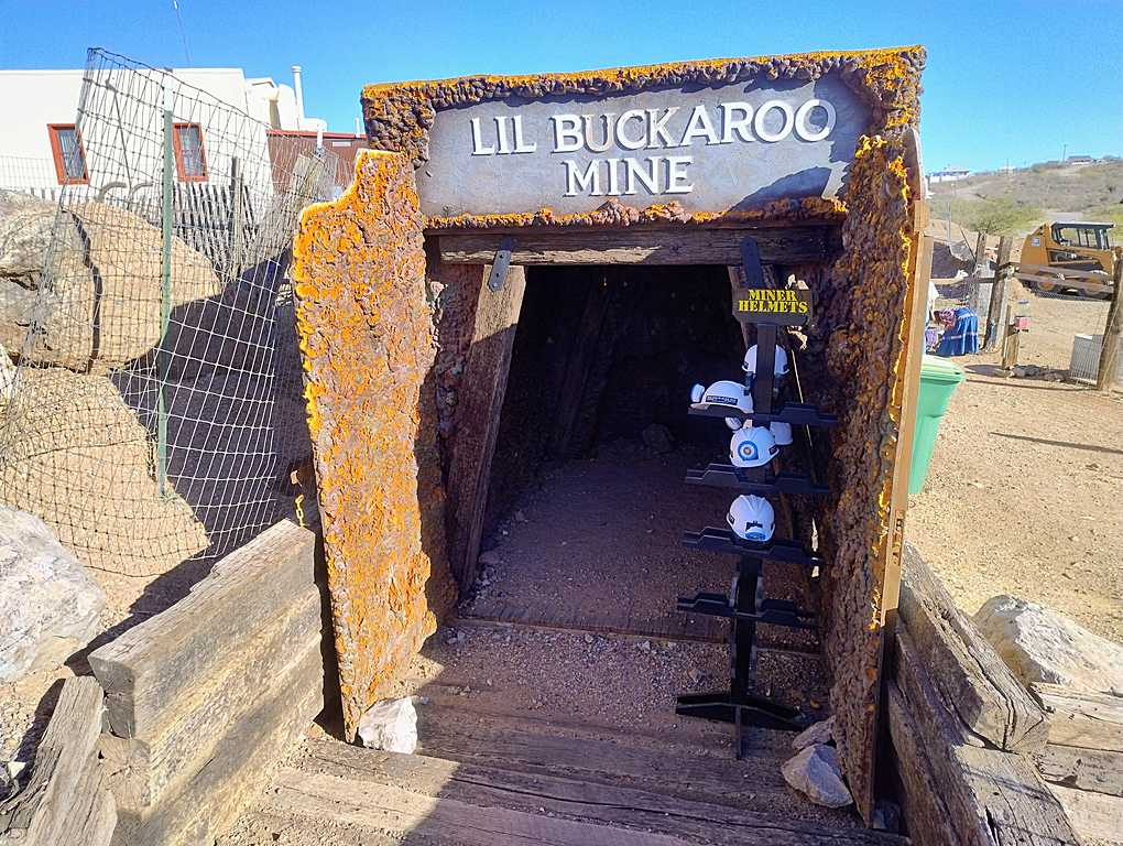 Entrance to the 'Lil Buckaroo Mine' with a rusted metal archway and a stand displaying miner helmets, set in a desert environment under a clear blue sky.