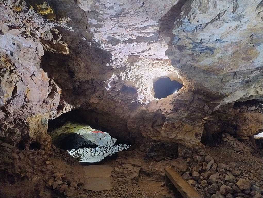 Underground mine chamber with rocky walls and ceiling, light streaming through openings, and piles of debris on the floor.