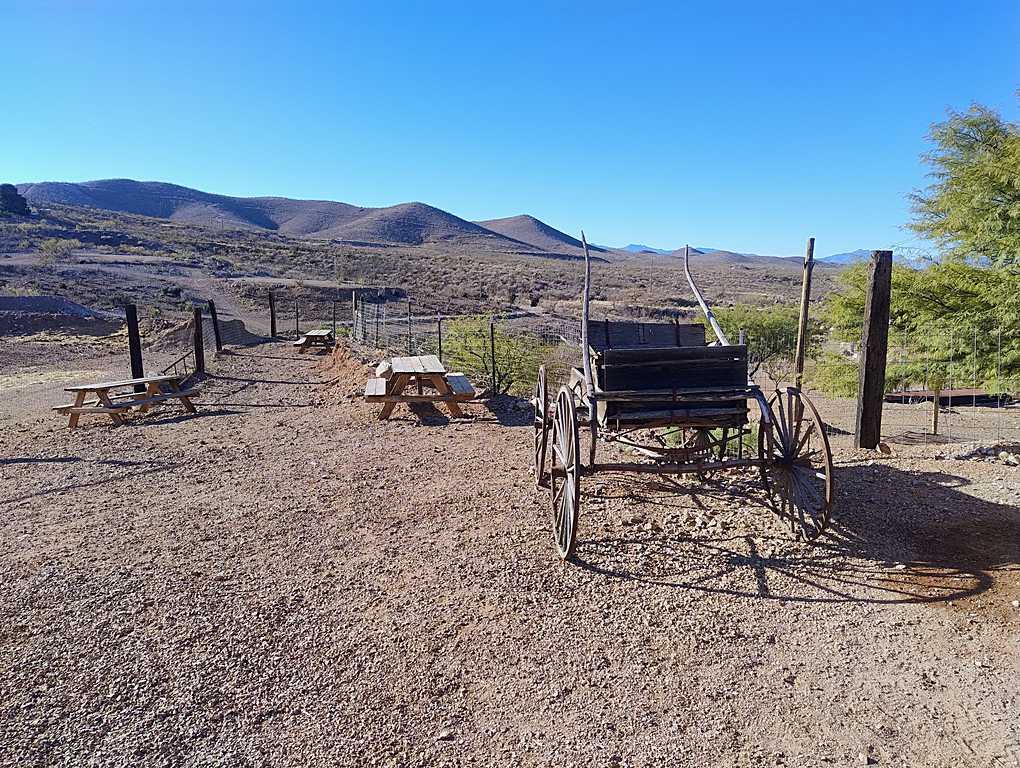 Outdoor area with a vintage wooden wagon on a gravel surface, picnic tables, and a backdrop of rolling hills under a clear blue sky.