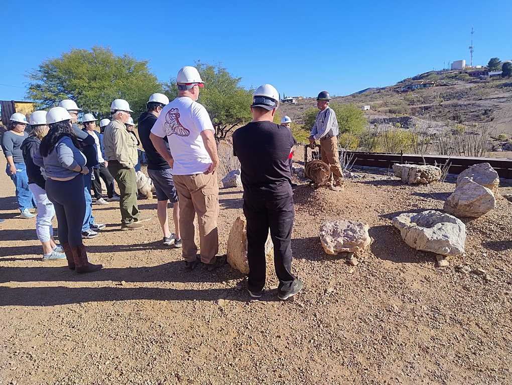 Group of people wearing hard hats gathered around a guide outdoors, surrounded by rocks and desert terrain under a clear blue sky.