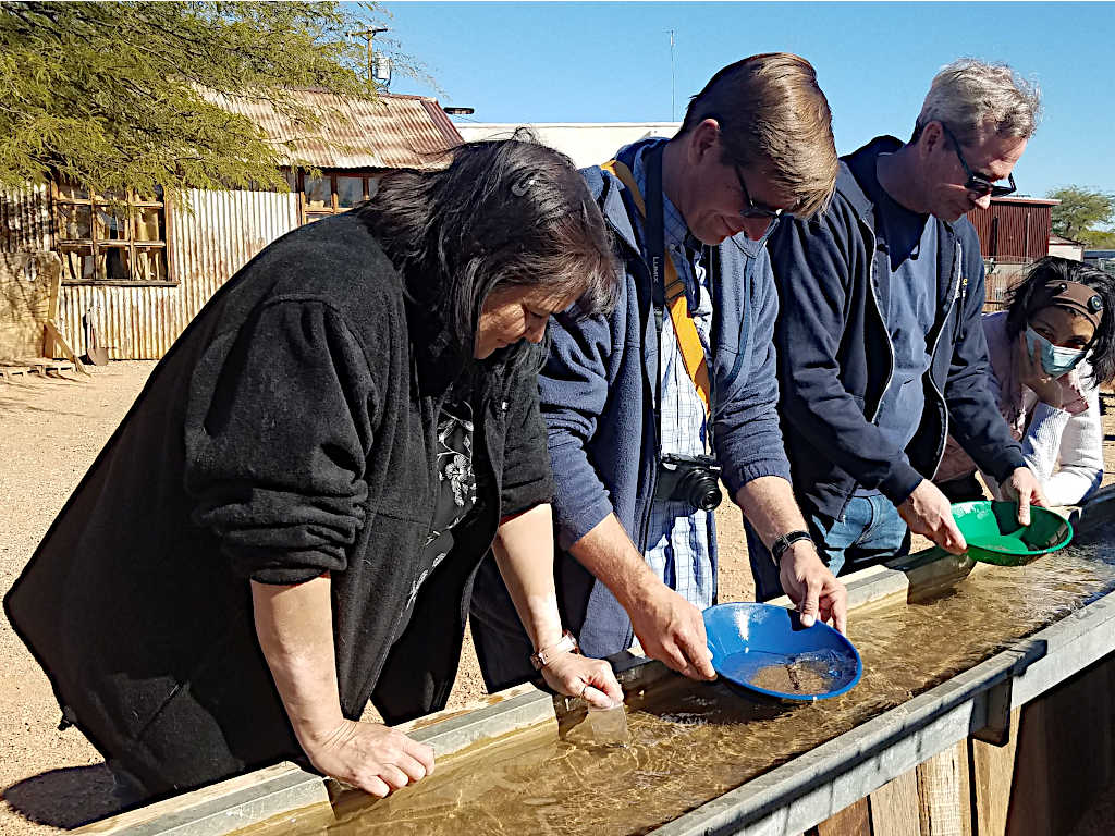 Group of people participating in a gold panning activity, using colorful pans over a wooden water trough, with rustic wooden buildings and desert trees in the background.