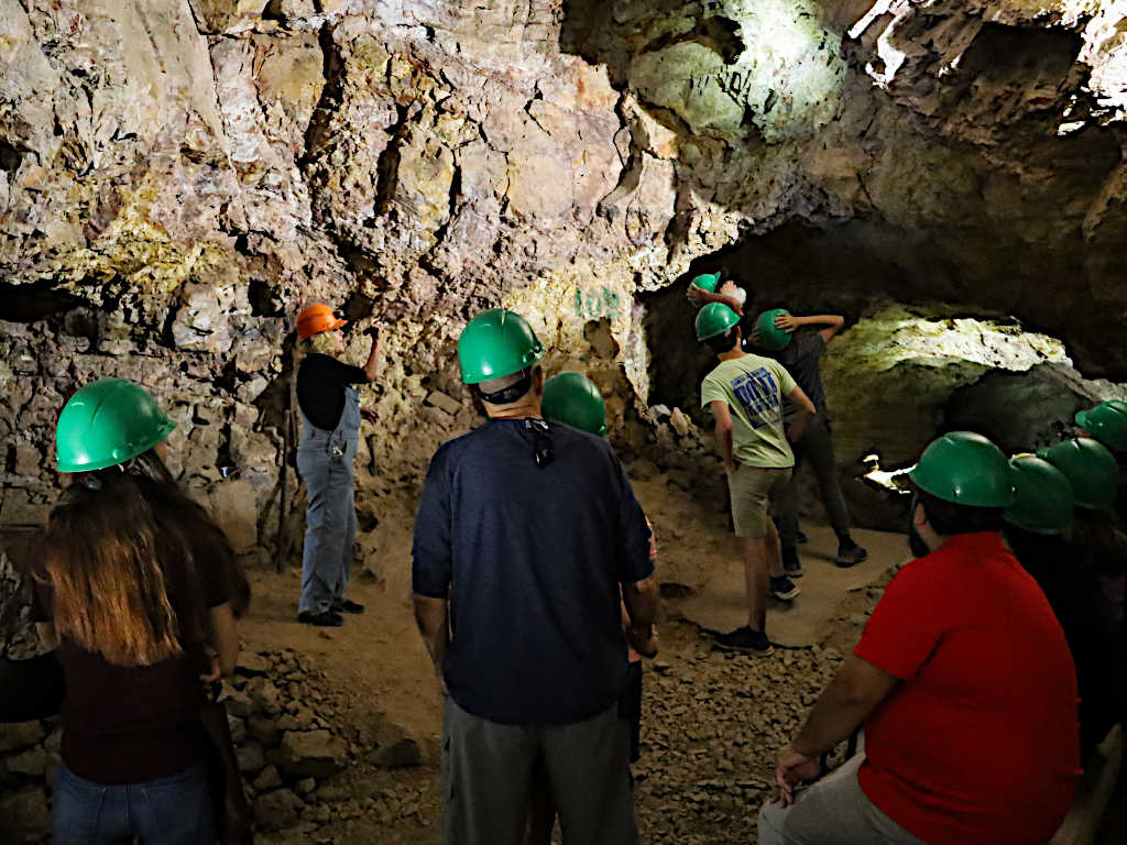 Tour guide wearing an orange hard hat explaining rock formations to a group of visitors wearing green hard hats inside an underground mine.