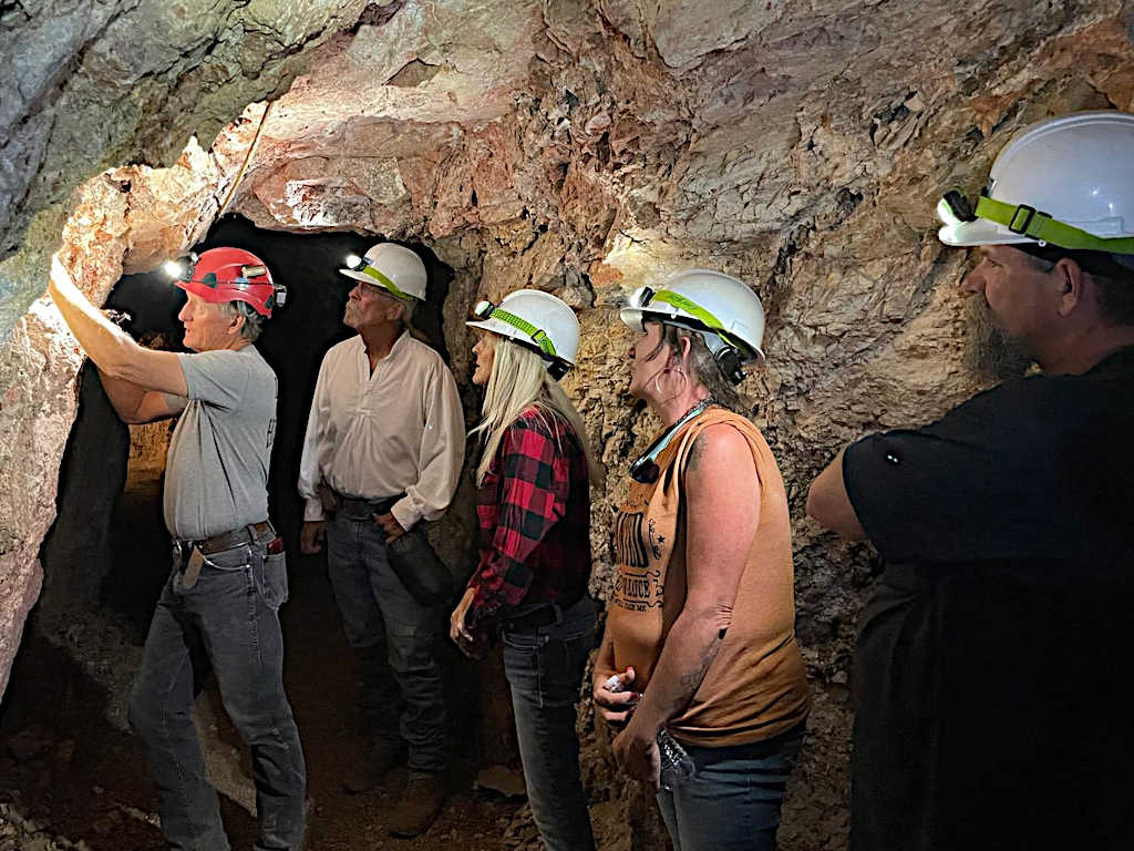 Tour guide wearing a red hard hat explaining mineral formations to a group of visitors inside a narrow underground mine passage, all wearing helmets with headlamps.