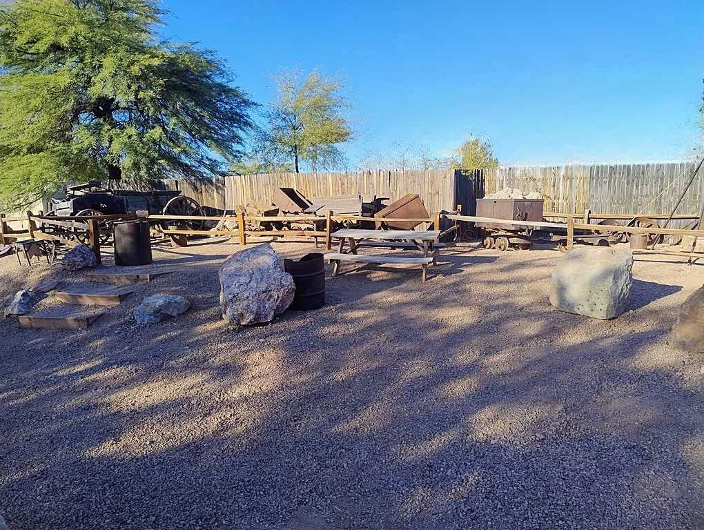 Outdoor display of historic mining equipment, wooden carts, and large rocks, set on gravel with a wooden fence and trees in the background.