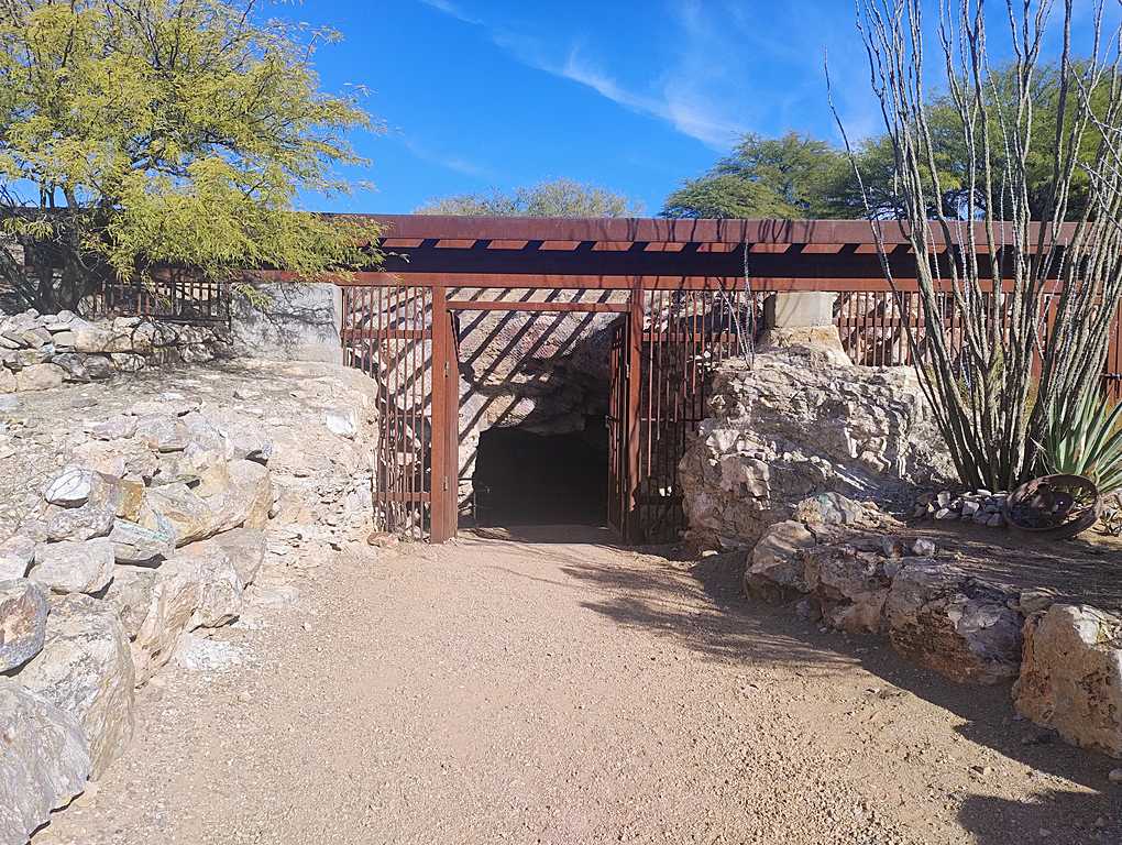 Entrance to an underground mine with a wooden walkway and metal handrails, surrounded by large rocks and rugged stone walls.