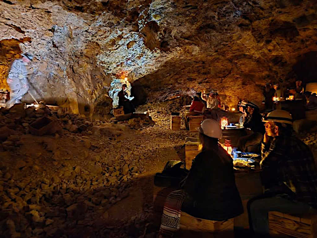 Underground mine chamber illuminated by lanterns showing visitors seated on wooden crates around barrel tables, watching a performer in period costume delivering a monologue in the atmospheric cave-like space with rocky walls and ceiling.