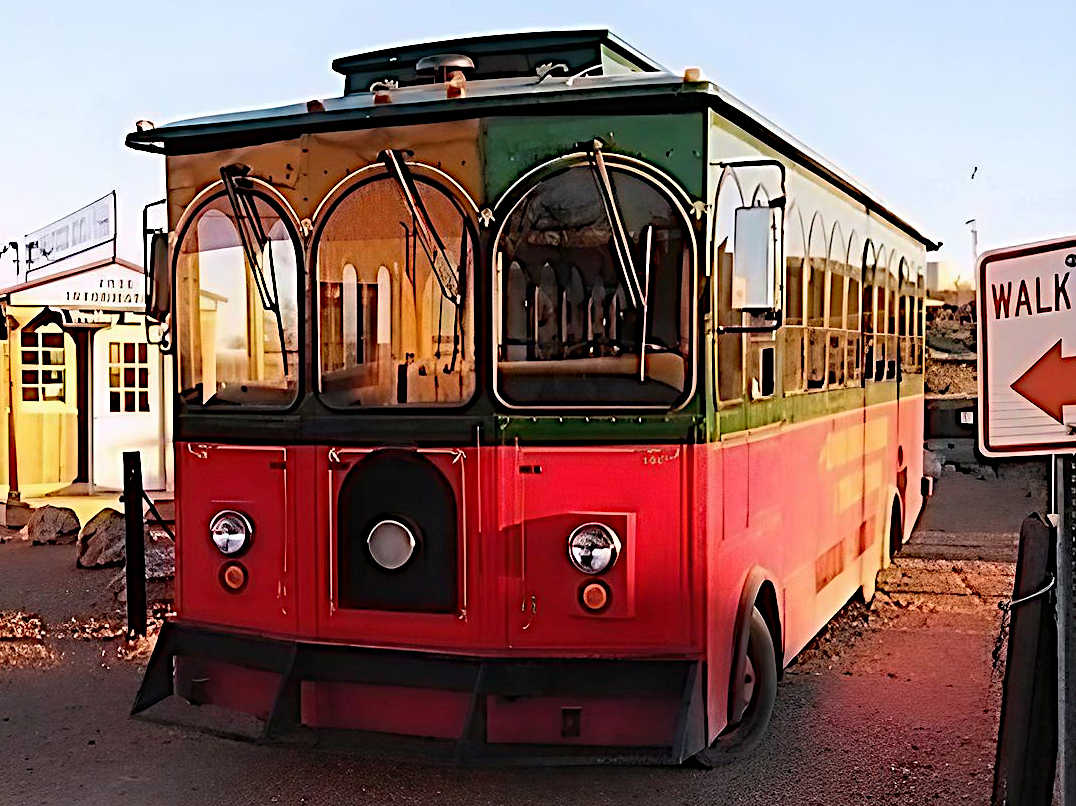 Front view of a red and green trolley bus parked near a 'WALK' sign with an old western-style building in the background, illuminated by warm sunlight.