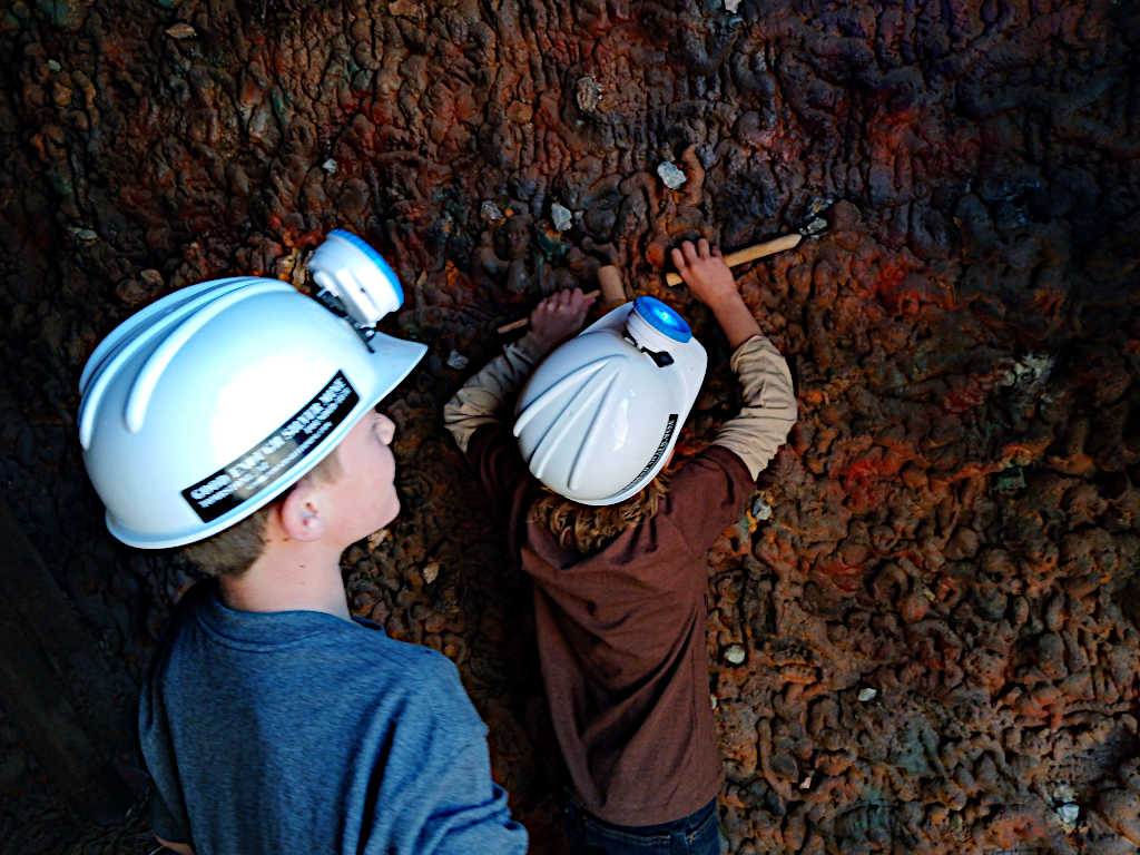 Two children wearing white hard hats with headlamps examine a colorful cave wall formation, with one child reaching up to touch the rock surface while another looks on. The wall displays rich orange, red, and blue mineral patterns.