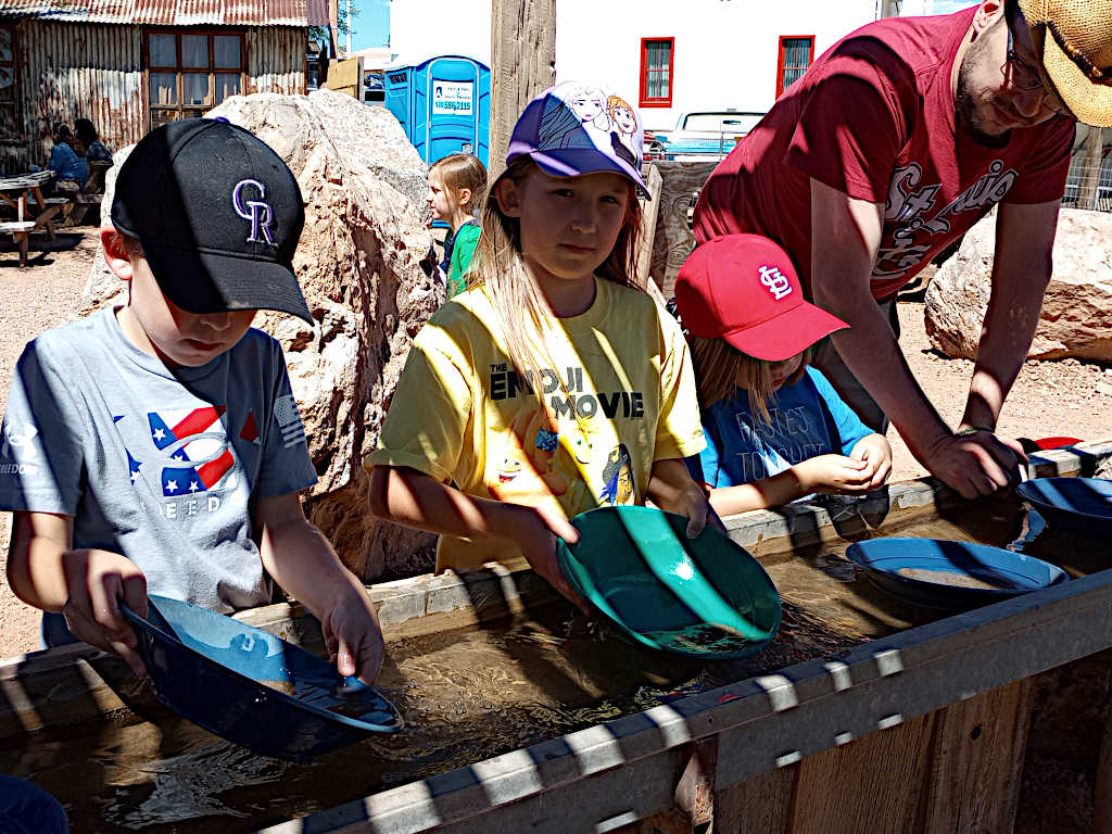 Group of young people participating in a gold panning activity, using colorful pans over a wooden water trough, with rustic wooden buildings in the background.