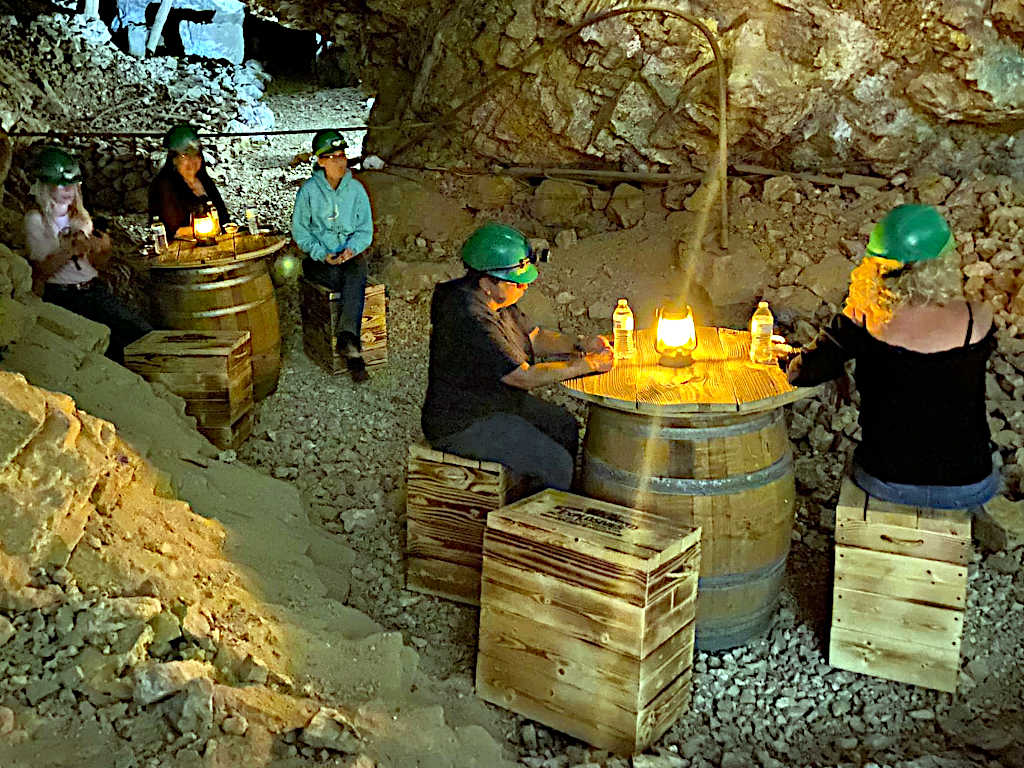 Visitors wearing green helmets seated on wooden crates around barrel tables with glowing lanterns, inside a rocky underground mine chamber.