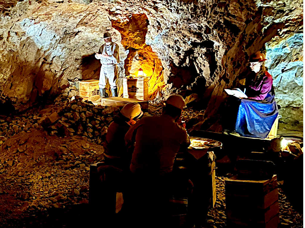 Performance inside an underground mine with actors in period costumes, one standing among wooden crates and another seated, illuminated by warm lighting.