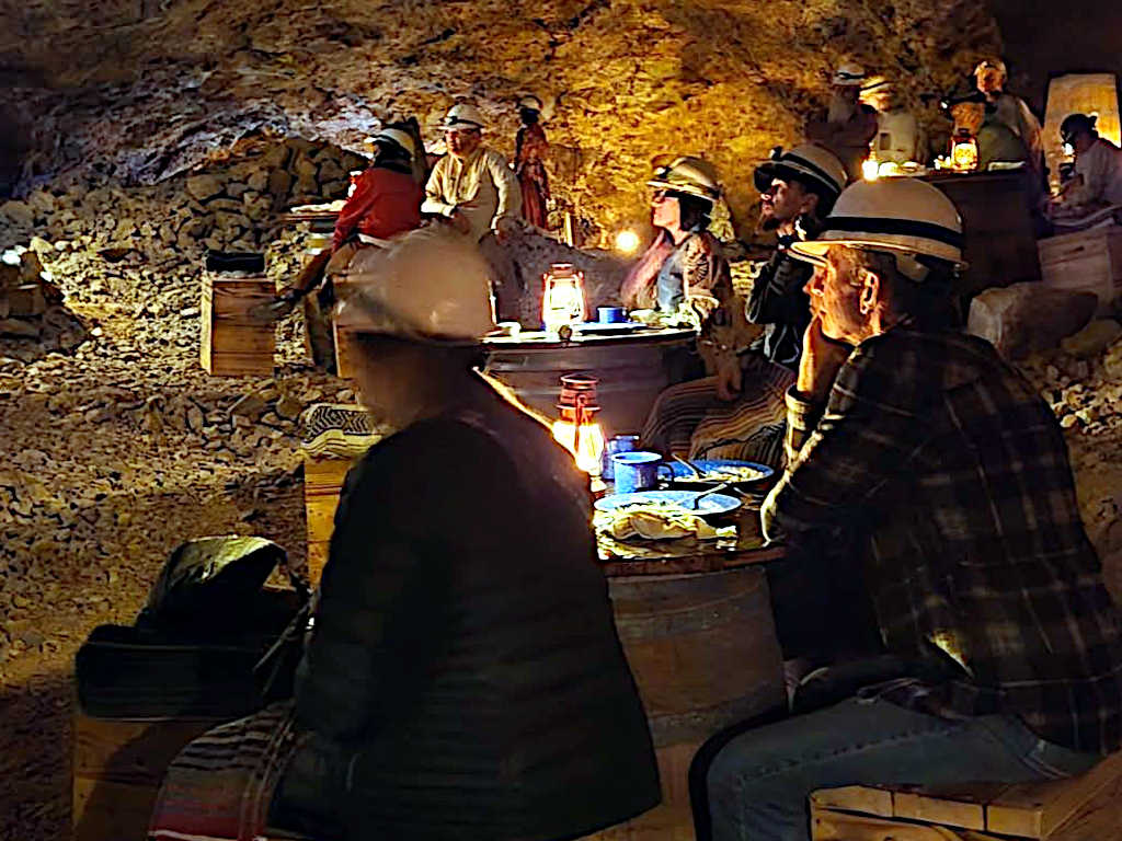 Audience seated at wooden tables in an underground mine, illuminated by lanterns, enjoying a performance in a cozy atmosphere.