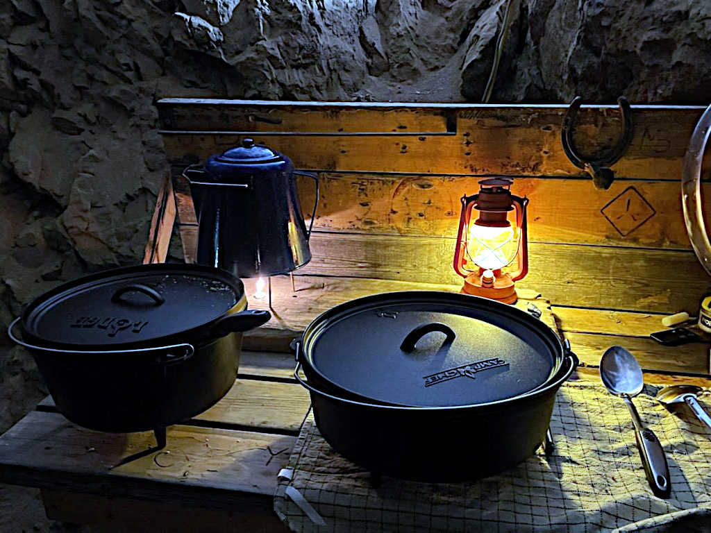 Rustic setup with cast iron pots, an enamel coffee pot, a glowing lantern, and utensils on a wooden table inside a rocky cave-like setting.