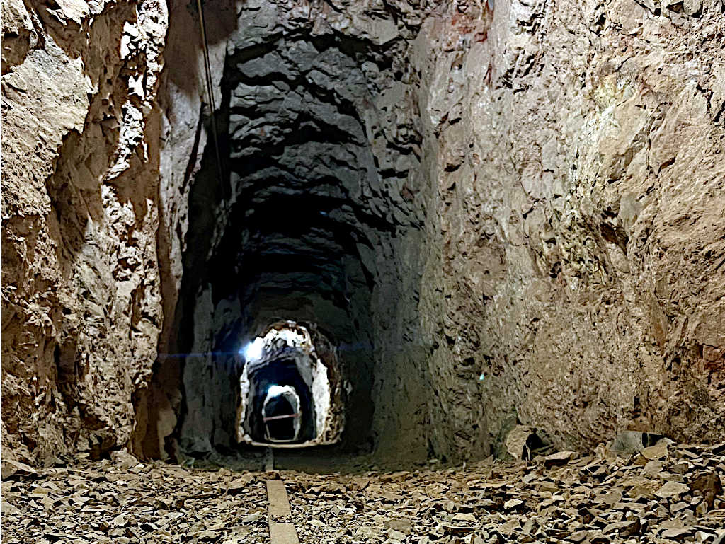 Long underground mine tunnel with rocky walls and a narrow track on the ground, illuminated by faint overhead lights in the distance.