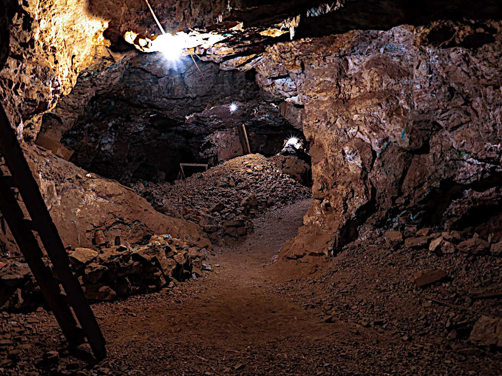 Underground mine chamber with rocky walls, scattered debris, and a ladder leaning against the wall, illuminated by overhead lights.