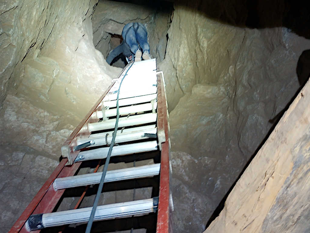 Interior view of a metal ladder with support rails extending upward inside a mine shaft, with rough stone walls visible and a person climbing at the top.