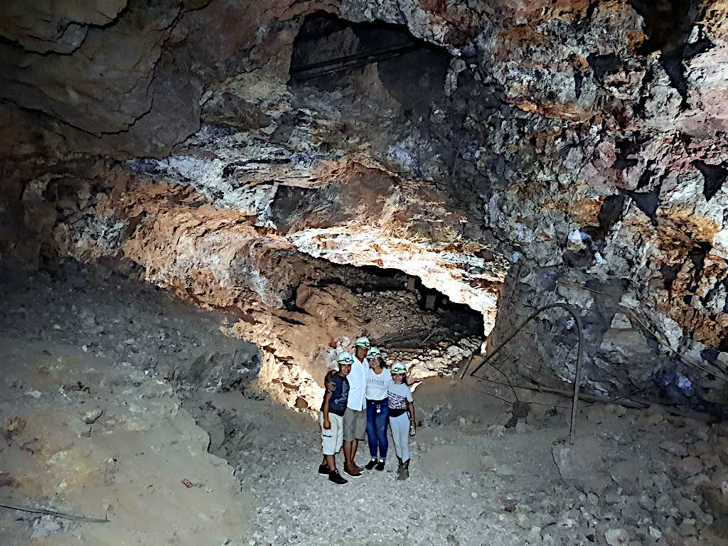 Group of visitors wearing helmets posing inside a large underground mine chamber with rugged rock formations and illuminated walls.