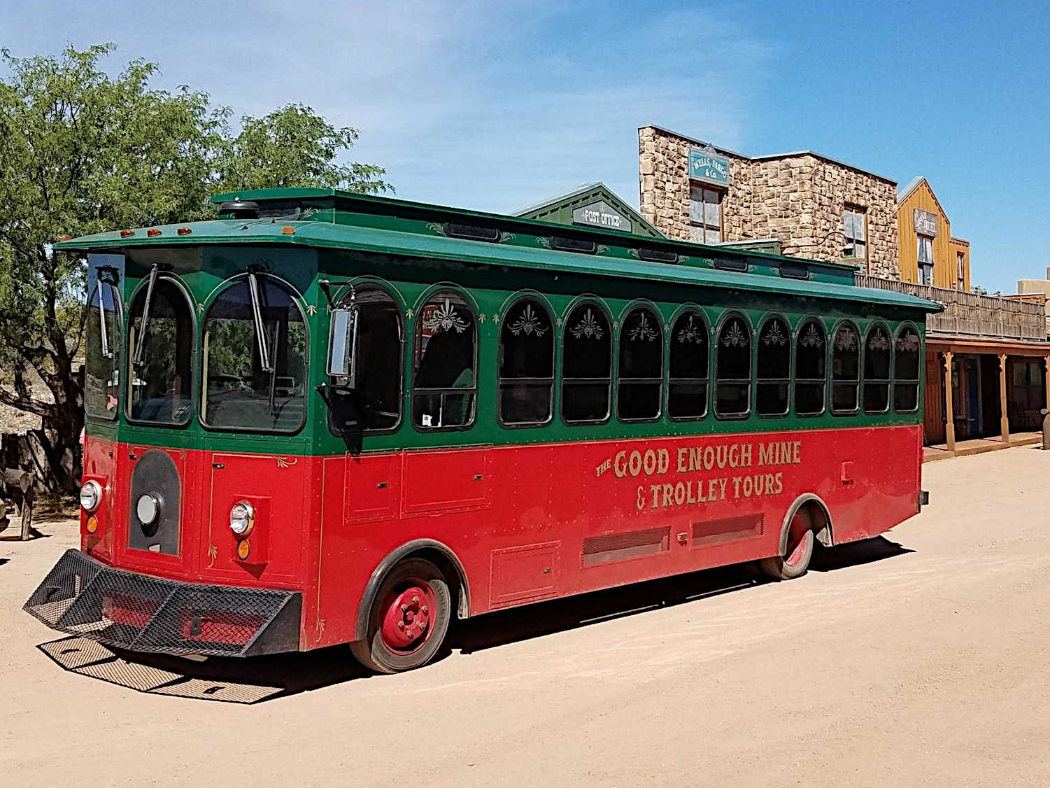 A red and green trolley labeled 'The Good Enough Mine & Trolley Tours,' parked on a dirt road with old western-style buildings and trees in the background.